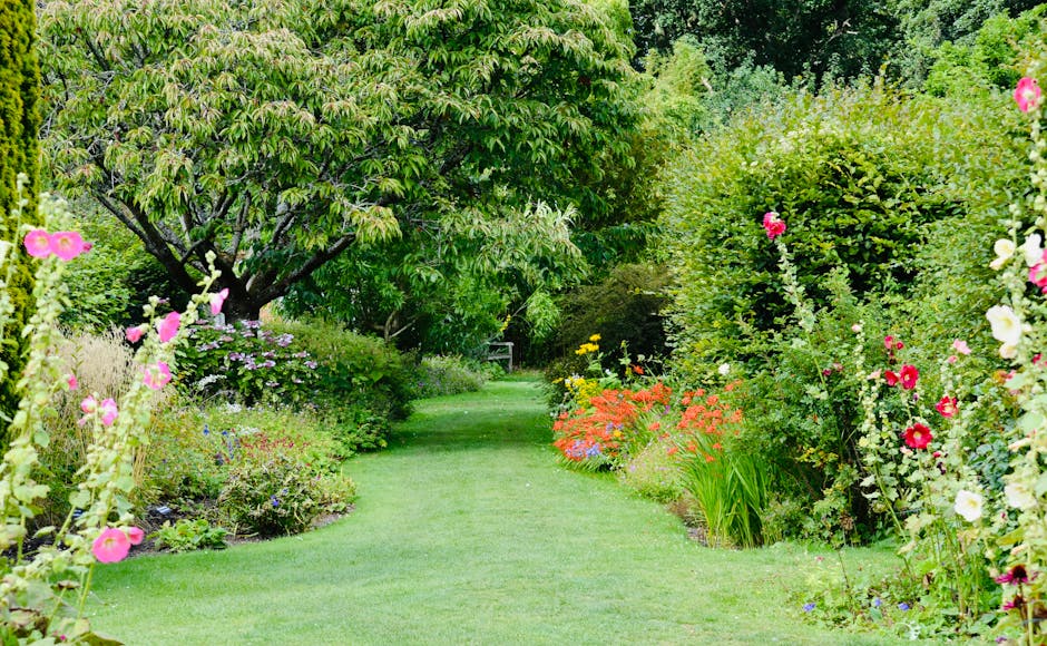A lush garden scene featuring a well-maintained grassy lawn bordered by an array of vibrant flowering plants and shrubs. On the left, pink hollyhocks and purple flowering bushes grow alongside a small