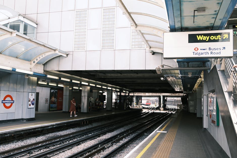 The image depicts the platform of Hammersmith station, with the railway tracks curving gently into the distance under a concrete overpass. The platform surface is made of dark paving with yellow tacti